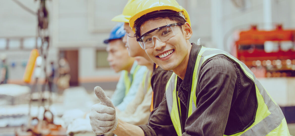 Young man in a hard hat, safety vest, and safety goggles giving a thumbs up and smiling at the camera